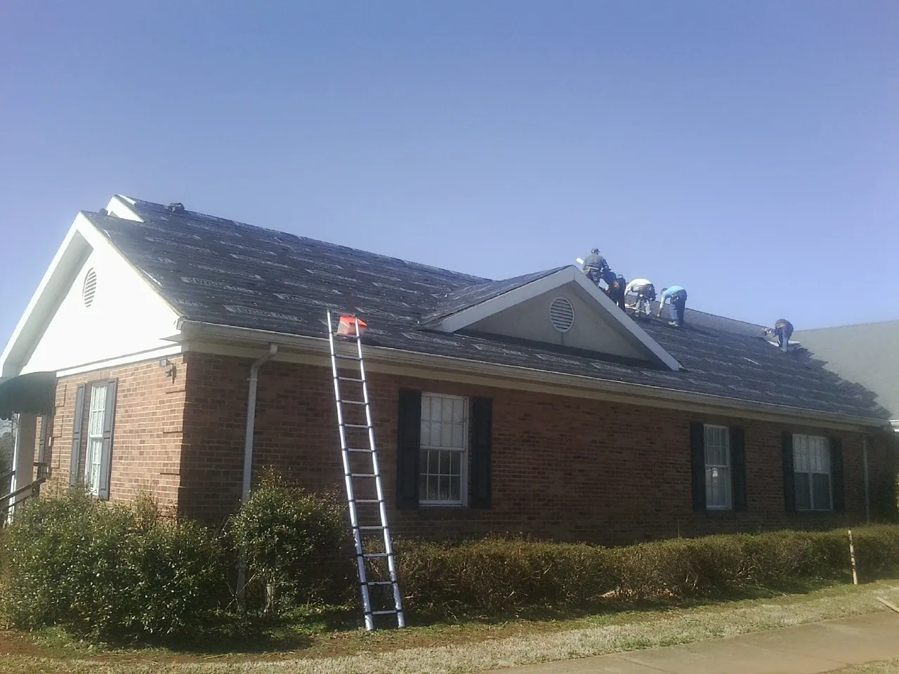 Crew tearing shingles off a brick ranch roof with a ladder set — Emergency Roof Repair work by our crew in Old Bethpage