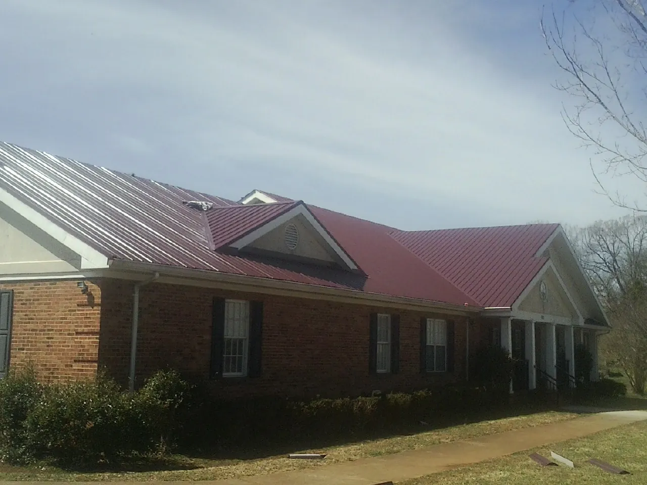 Red metal roof with gabled entries on a long brick building, from a Hail Damage Roof Repair job in Old Bethpage
