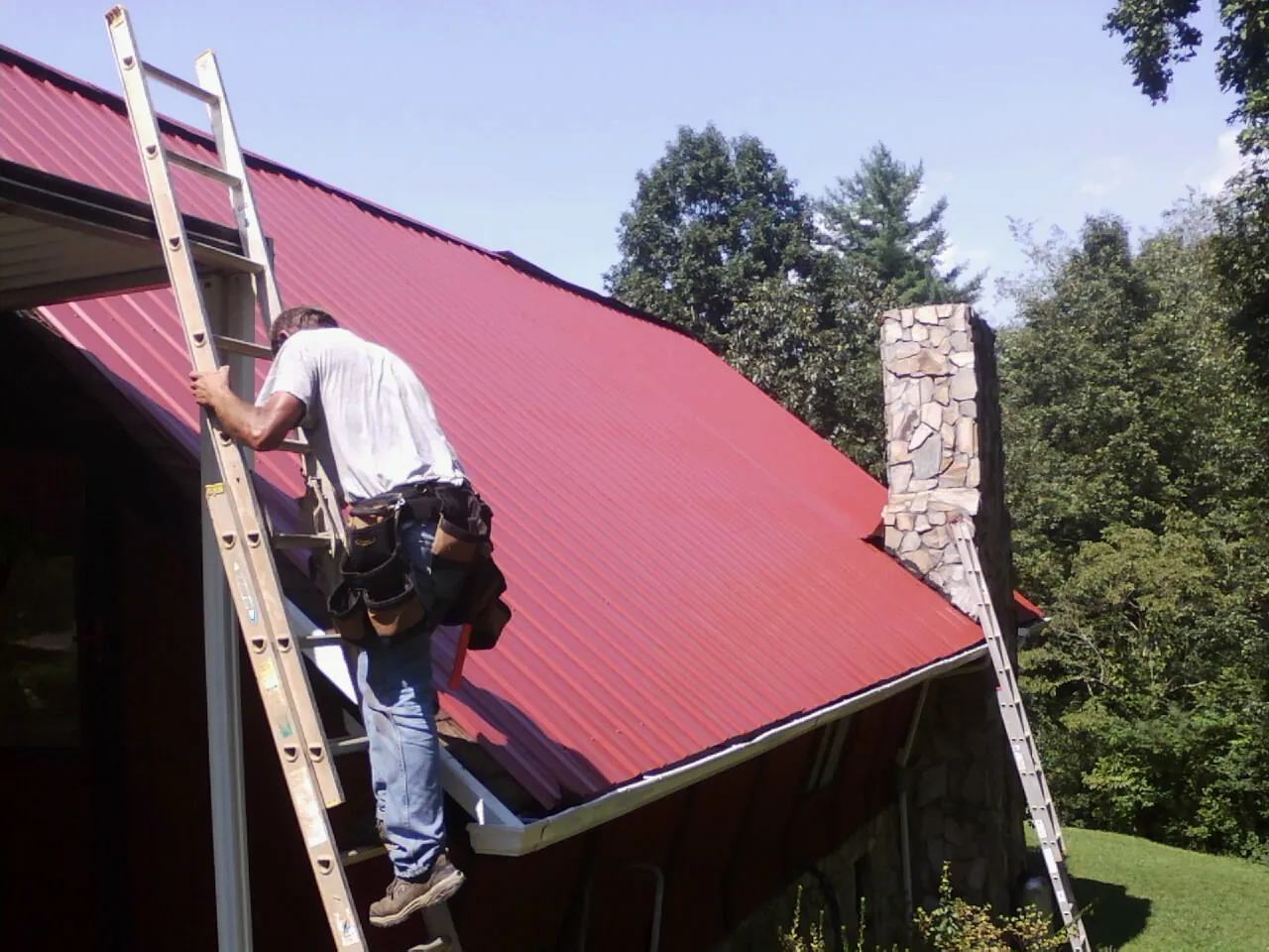 Roofer on a ladder beside a red metal roof and stone chimney — Soffit & Fascia Repair by Roofing Brothers in Old Bethpage