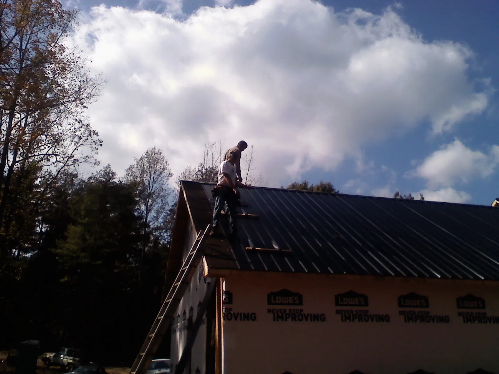 Roofer fastening dark metal panels high against a cloudy sky, from a Roof Inspection job in Old Bethpage